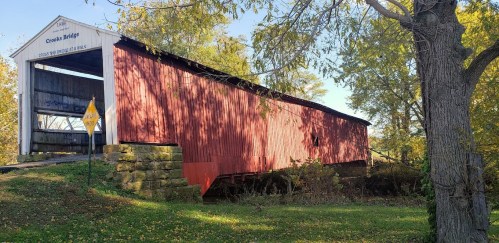 A red covered bridge stands surrounded by trees, with a sign nearby and sunlight filtering through the leaves.