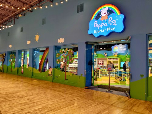 Entrance to Peppa Pig World of Play, featuring colorful decor, trees, and a gift shop sign. Bright and inviting atmosphere.