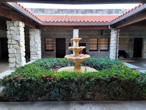 A stone fountain surrounded by greenery in a courtyard with rustic architecture and a tiled roof.