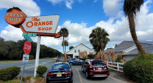 A roadside view of The Orange Shop sign with palm trees and parked cars under a blue sky with clouds.