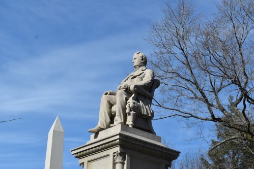 A seated statue of a man on a pedestal, with a tall obelisk and bare trees against a blue sky in the background.