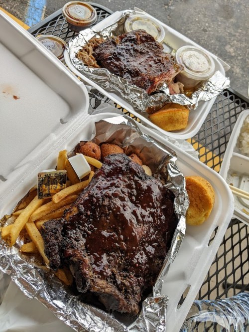 Two takeout containers with barbecue ribs, fries, cornbread, and dipping sauces on a table.