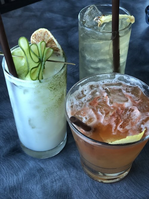 Three colorful cocktails on a table: a green drink with cucumber, a clear drink with ice, and a brown drink with a lemon slice.