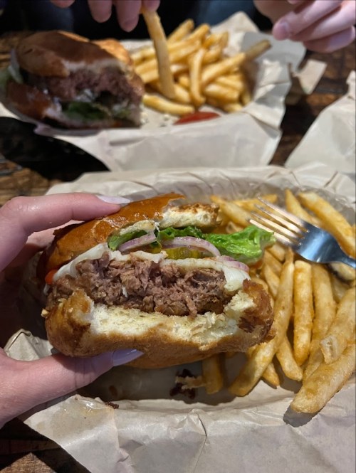 A close-up of a hand holding a burger with lettuce and onion, alongside a plate of fries in the background.