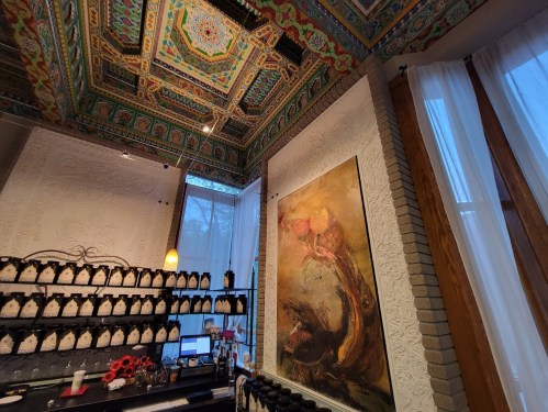 A colorful, intricately designed ceiling above a bar area with a large painting and shelves of bottles.