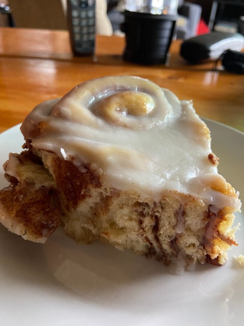 A slice of cinnamon roll topped with white icing, resting on a plate with a wooden table in the background.