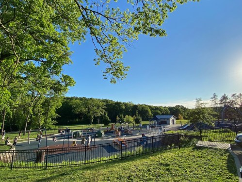 A sunny playground scene with children playing, surrounded by trees and a fence, under a clear blue sky.