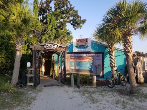 A colorful beachside restaurant entrance with palm trees and an "OPEN" sign, surrounded by sand and greenery.