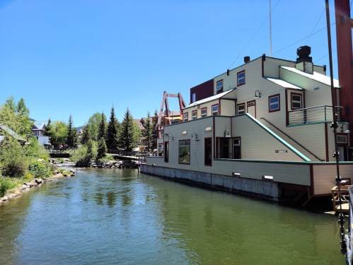 A charming building by a calm river, surrounded by trees and blue skies, with a bridge in the background.