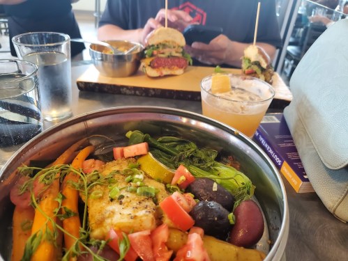 A colorful bowl of grilled fish and vegetables, with a cocktail and burgers in the background at a dining table.