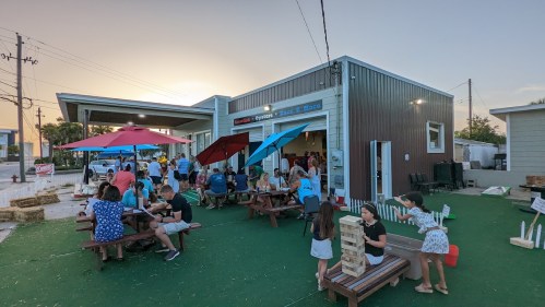 A lively outdoor scene at a restaurant with people dining, playing games, and colorful umbrellas at sunset.
