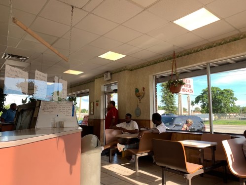 Interior of a casual restaurant with customers seated, large windows, and a menu board visible. Bright, sunny atmosphere.