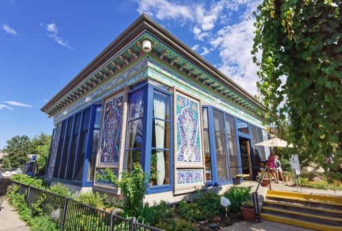 Colorful building with intricate tile work, large windows, and a patio, surrounded by greenery and blue skies.