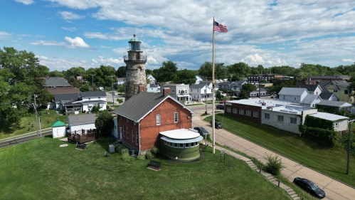 Aerial view of a small town featuring a lighthouse, flagpole, and various buildings under a partly cloudy sky.