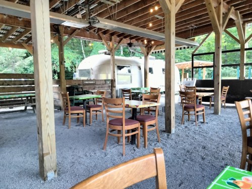 A rustic outdoor dining area with wooden tables and chairs, surrounded by trees and an Airstream trailer in the background.