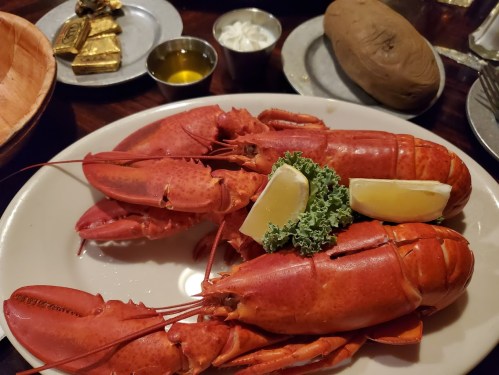 A plate with two cooked lobsters, garnished with lemon wedges and parsley, alongside a baked potato and dipping sauces.