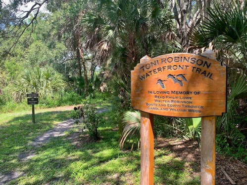 Wooden sign for Toni Robinson Waterfront Trail, surrounded by greenery, with a path leading into the natural area.