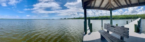 A panoramic view of a calm lake with a wooden dock and a shaded seating area under a gazebo. Bright blue sky and clouds.