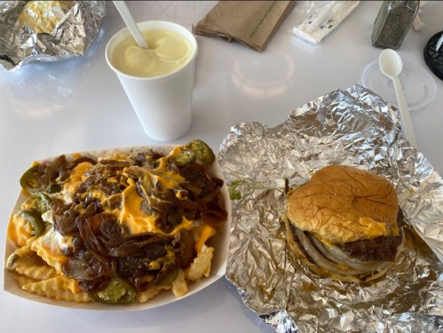 A plate of loaded fries topped with cheese and peppers, alongside a wrapped burger and a cup of drink.