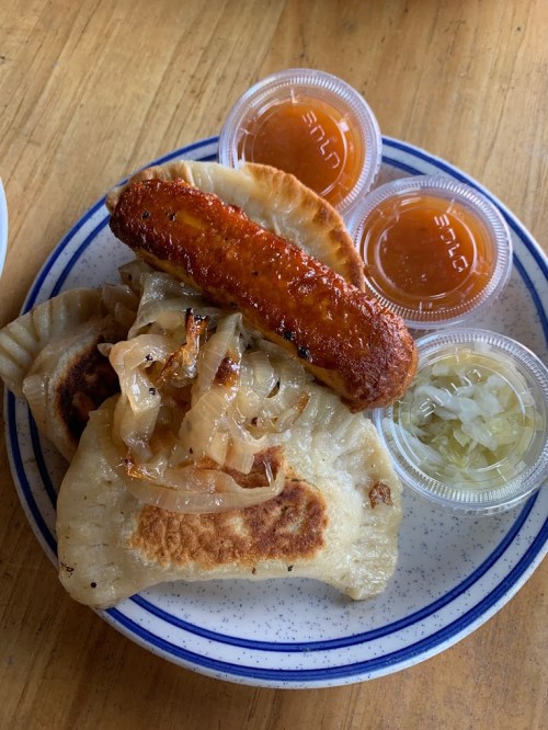 A plate of pierogi topped with sautéed onions, a sausage, and two containers of sauce, alongside pickled cabbage.