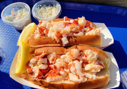 Two lobster rolls filled with chunks of lobster meat, served with a lemon wedge and two cups of coleslaw on a blue tray.