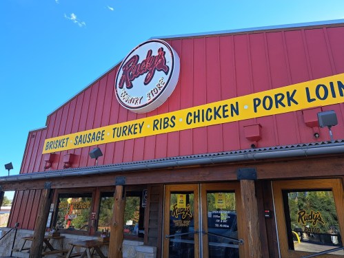 Exterior of Rudy's Country Store with a red facade and signage for various meats like brisket, ribs, and chicken.
