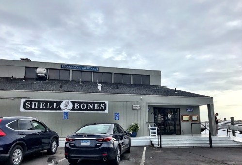Exterior of Shell Bones restaurant with a cloudy sky, featuring a sign and parked cars in front.