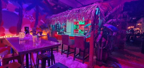 A vibrant bar with tropical decor, colorful lighting, and a thatched roof, surrounded by palm trees and a lively atmosphere.