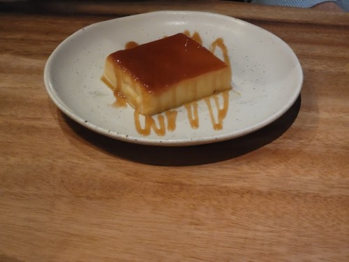 A smooth, rectangular flan topped with caramel sauce on a white plate, resting on a wooden surface.