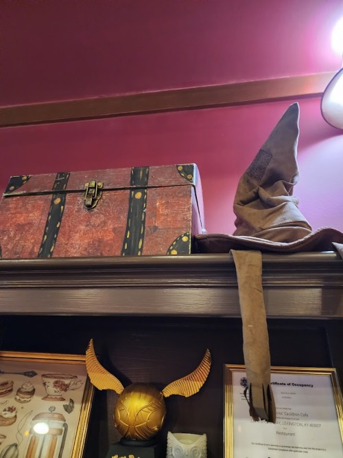 A wooden chest and a pointed hat sit atop a shelf against a pink wall, with decorative items below.
