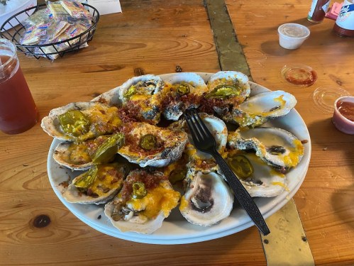 A plate of baked oysters topped with cheese, jalapeños, and bacon, served on a wooden table with sauces and a drink.