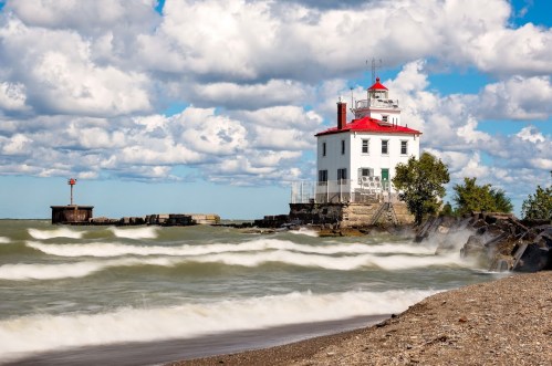 A white lighthouse with a red roof stands by a rocky shore, with waves crashing and clouds in a blue sky.