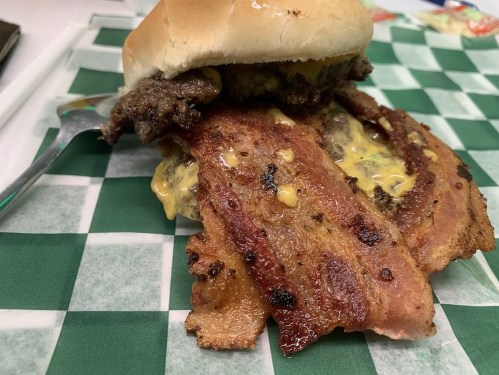 A close-up of a sandwich with a beef patty, melted cheese, and crispy bacon on a soft bun, resting on a green checkered tablecloth.