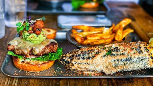A gourmet burger with cheese, guacamole, and greens, served with fries and a grilled corn on a metal tray.