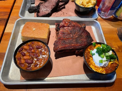 A metal tray with barbecue brisket, cornbread, baked beans, and chili topped with cheese and green onions.