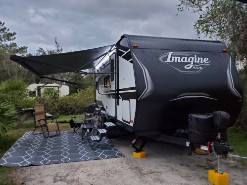 A black and white travel trailer with an awning set up at a campsite, surrounded by greenery and outdoor furniture.