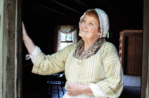 A smiling older woman in historical attire stands in a doorway, looking thoughtfully outside.
