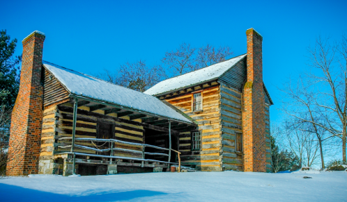 A historic log cabin with snow covering the ground, featuring two chimneys and a porch, under a clear blue sky.