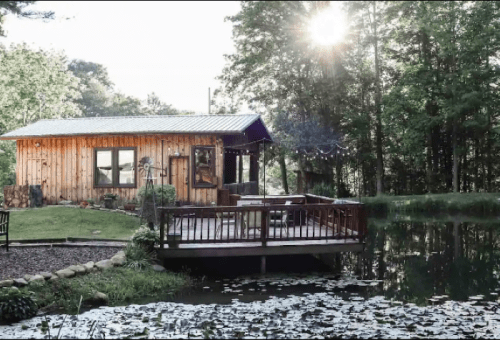 A cozy wooden cabin by a serene pond, surrounded by trees and sunlight filtering through the leaves.