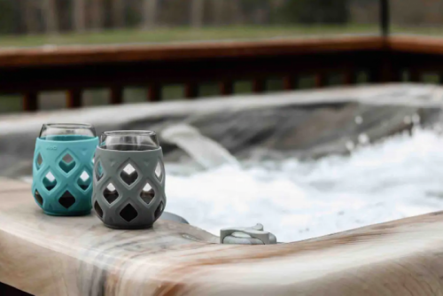 Two decorative glasses sit on the edge of a hot tub, with bubbling water and a wooden deck in the background.