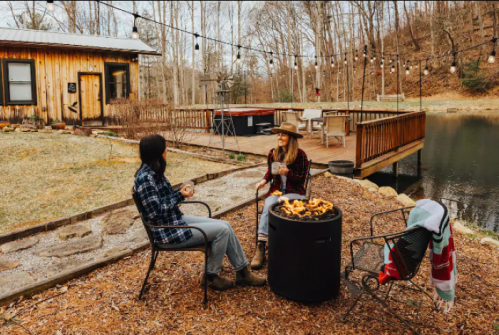 Two people sit by a fire pit in a cozy outdoor setting, surrounded by trees and a pond, enjoying a relaxed moment.
