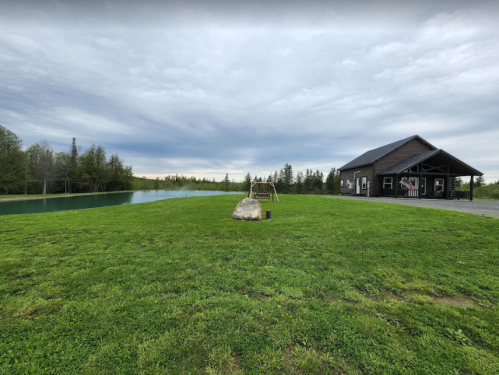 A serene landscape featuring a black cabin by a pond, surrounded by green grass and trees under a cloudy sky.