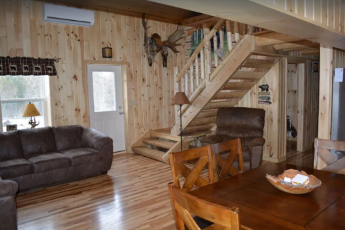 Cozy wooden interior of a cabin featuring a living area, dining table, and staircase. Natural light from the door.