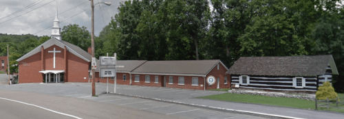 A brick church with a cross and a log cabin nearby, set along a road surrounded by trees.