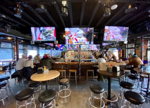 A lively bar interior with multiple TVs showing sports, patrons seated at the bar, and a well-stocked drink counter.