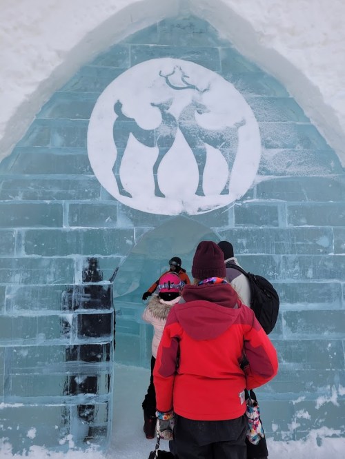 People entering an ice structure with a reindeer emblem on the icy archway. Snow surrounds the entrance.