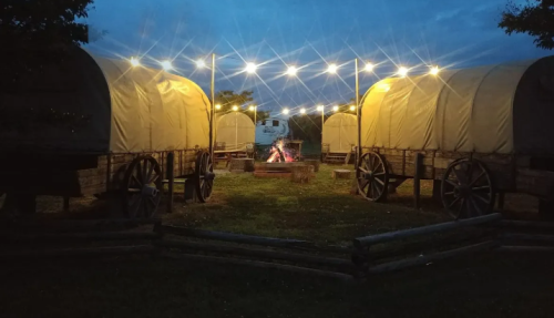 Two covered wagons are illuminated by string lights at dusk, with a campfire in the center and a rustic wooden fence.