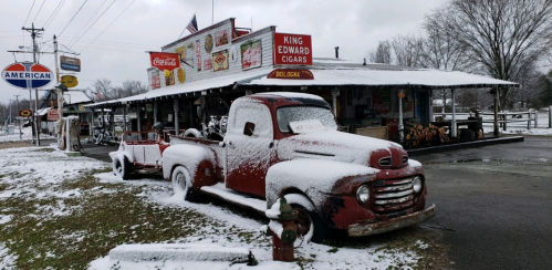 A vintage red truck covered in snow parked outside a rustic store with signs, surrounded by a winter landscape.