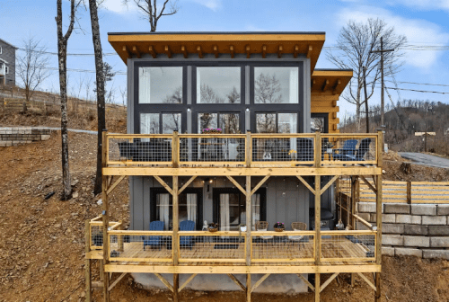 Modern two-story house with large windows, wooden deck, and a hillside setting, surrounded by trees.