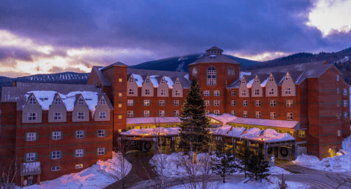 A large red hotel with snow-covered roofs, surrounded by trees and mountains, under a twilight sky.